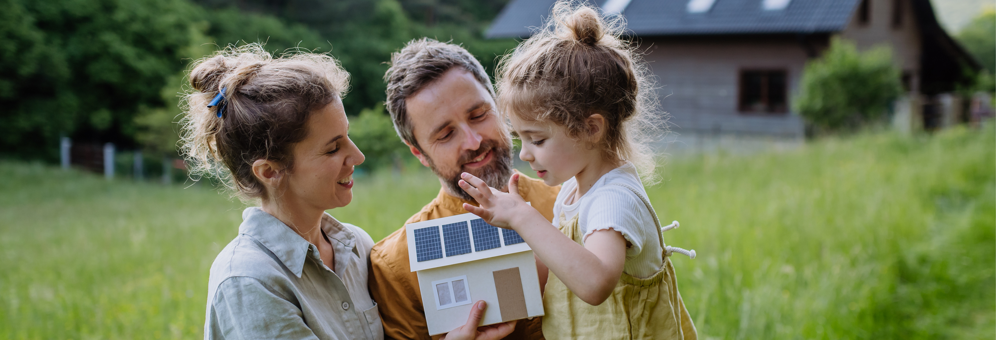Eine Personengruppe steht auf einer Wiese und hält ein Modellhaus mit Solarpaneelen, im Hintergrund ein Haus mit Solardach